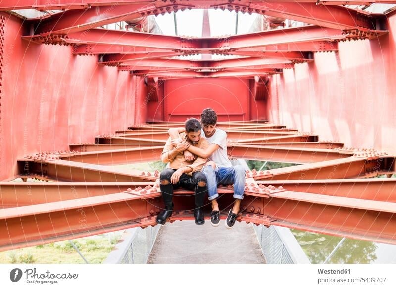 Young gay couple in love sitting on steel girder of a footbridge looking down twosomes partnership couples foot bridges Steel Girders gay men gay man