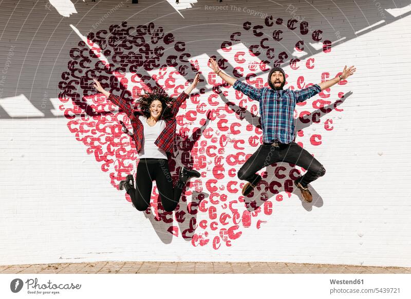 Happy young couple jumping in front of a brick wall with a heart Tarragona mid-air midair mid air bricks togetherness Arms outstretched outstretched arms