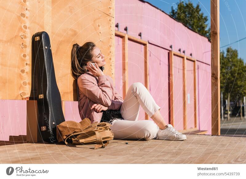 Smiling young woman sitting on platform next to guitar case listening to music Seated females women Guitar Case smiling smile hearing Adults grown-ups grownups