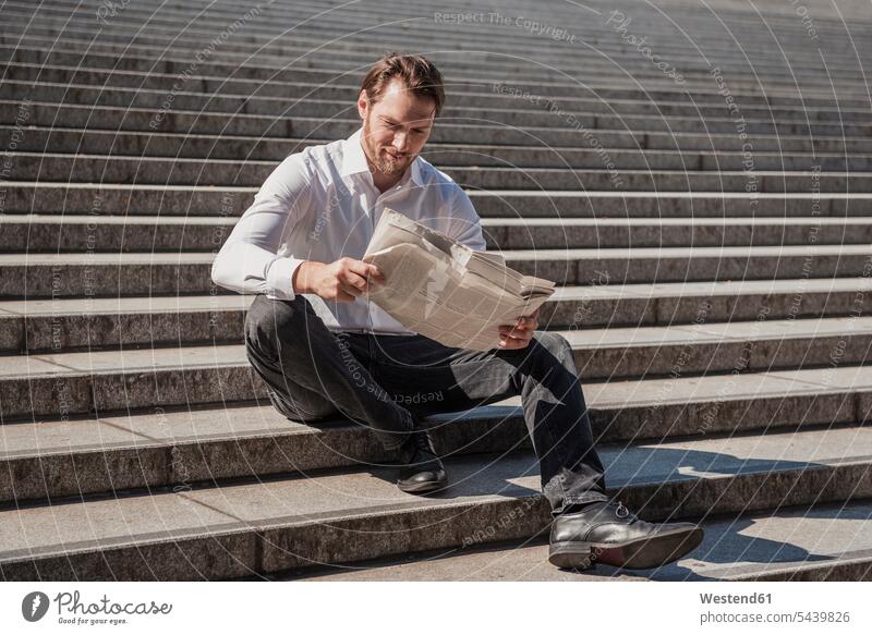 Smiling businessman sitting on stairs reading newspaper Seated Businessman Business man Businessmen Business men stairway smiling smile newspapers