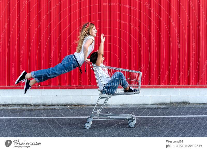 Sisters with shopping cart in front of red wall pants Trouser Denim Jeans T- Shirt t-shirts tee-shirt shopping trolley buy smile Seated sit play Ardor Ardour