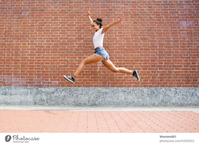 Exuberant young woman jumping in front of a brick wall Fun having fun funny happiness happy Leaping females women brick walls jumps Adults grown-ups grownups