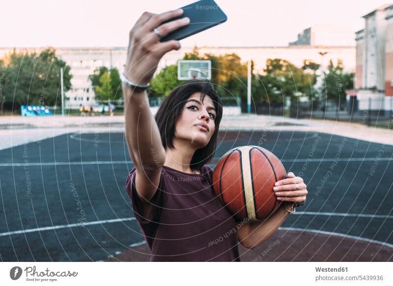 Young woman with basketball taking a selfie on outdoor court females women Selfie Selfies basketballs sports field sports fields Adults grown-ups grownups adult