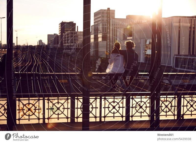 Germany, Munich, Young couple sitting on bridge, enjoying sunset sunsets sundown embracing embrace Embracement hug hugging Future Seated twosomes partnership