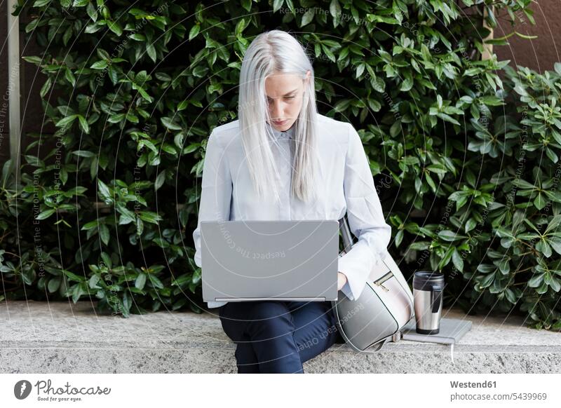 Young businesswoman sitting on a wall in the city using laptop Laptop Computers laptops notebook businesswomen business woman business women Seated females