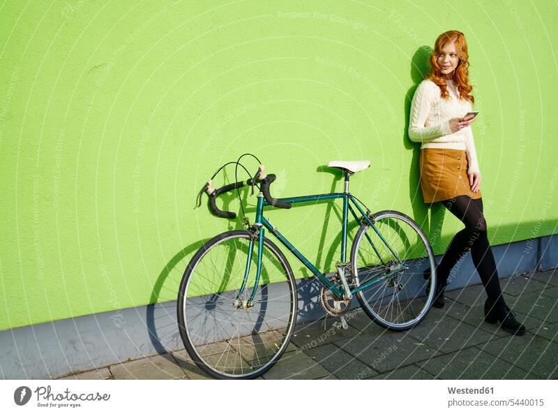 Young woman with cell phone lenaing against a green wall next to bicycle bikes bicycles mobile phone mobiles mobile phones Cellphone cell phones females women