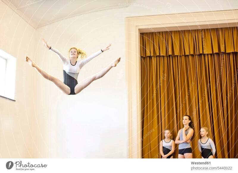 Girl in gymnastics outfit doing aerial jump with three girls watching team teams Arms outstretched arms out arm out arm outstretched gymnasium gyms gymnasiums