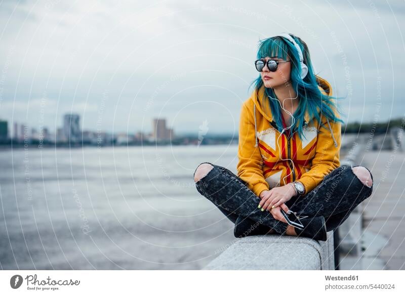 Young woman with dyed blue hair sitting on a wall listening music with headphones and smartphone females women Seated Smartphone iPhone Smartphones hearing