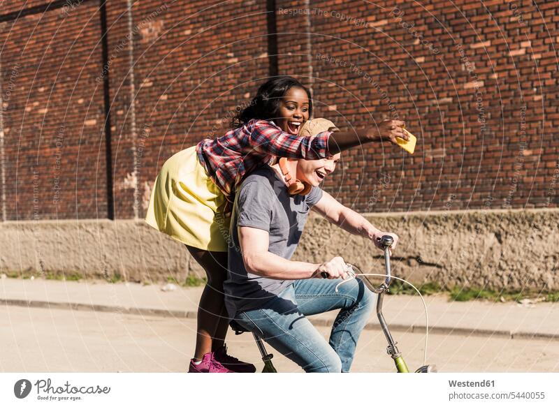Young couple riding bicycle in the street, woman standing on rack, taking selfies riding bike bike riding cycling bicycling pedaling Selfie Selfies mobile phone