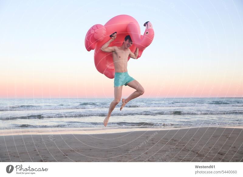 Laughing young man jumping in the air on the beach with inflatable pink flamingo beaches Leaping men males jumps Adults grown-ups grownups adult people persons
