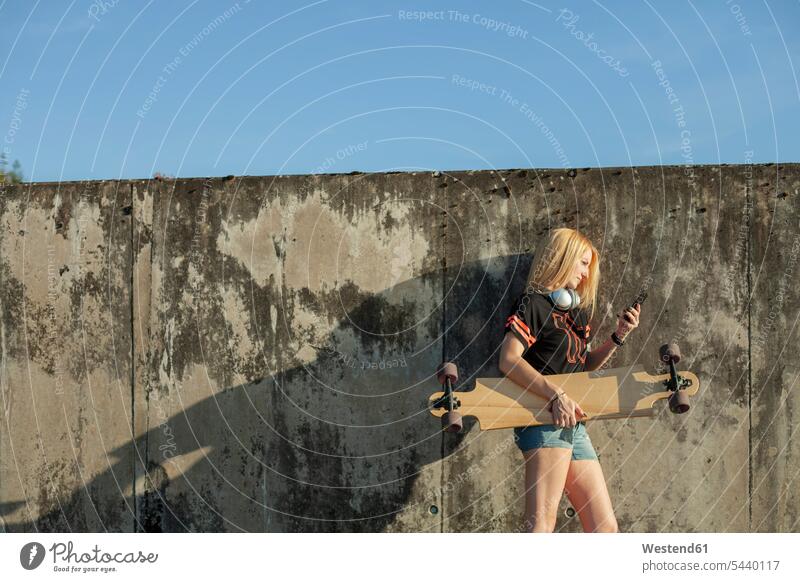 Blond woman with longboard standing in front of wall looking at smartphone Smartphone iPhone Smartphones walls females women Longboard view seeing viewing