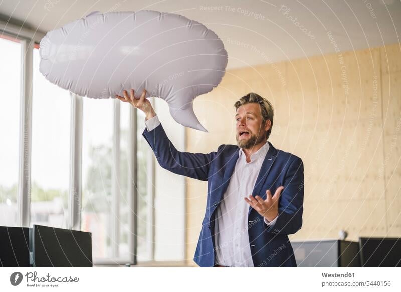 Businessman standing in office, holding inflatable speech bubble blow-up Business man Businessmen Business men offices office room office rooms speech balloon