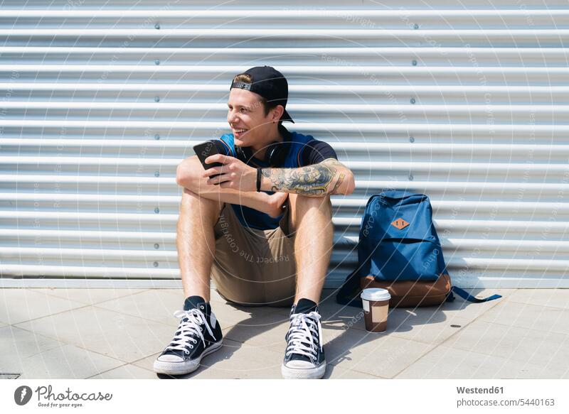 Smiling young man with smartphone, backpack and coffee to go sitting on the ground waiting men males Adults grown-ups grownups adult people persons human being
