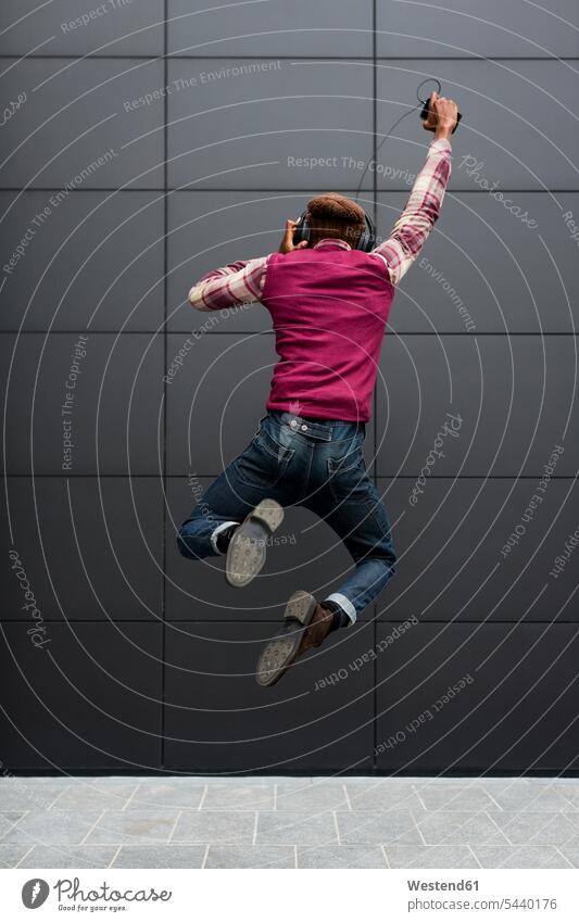 Back view of happy man jumping in the air while listening music with headphones and cell phone Leaping men males headset jumps Adults grown-ups grownups adult