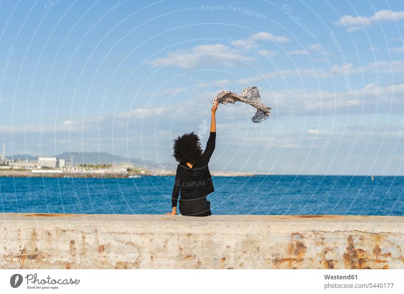 Spain, Barcelona, back view of woman dressed in black sitting on wall waving with scarf Seated females women walls Neck Scarves scarves Neck Scarf wave Adults