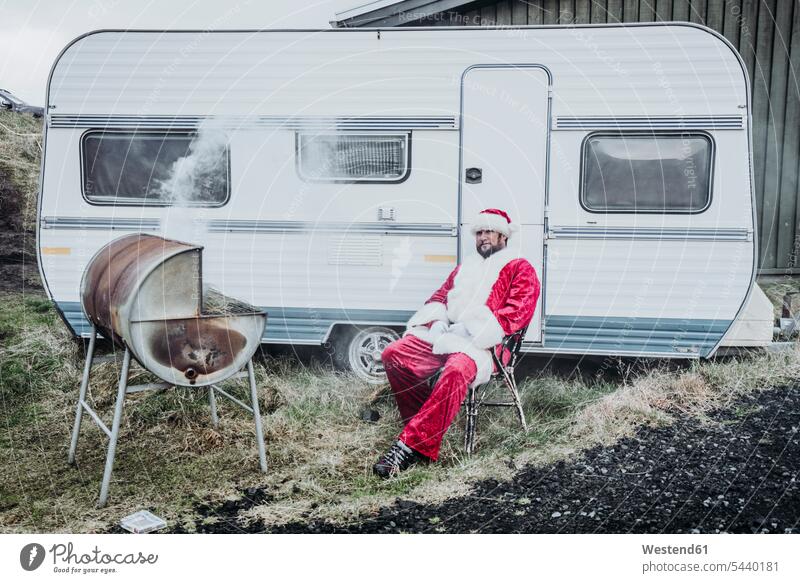 Iceland, Santa Claus sitting in front of caravan barbecueing Father Christmas Travel Trailers Camper Trailer mobile home Barbecuing grilling Seated X-Mas yule