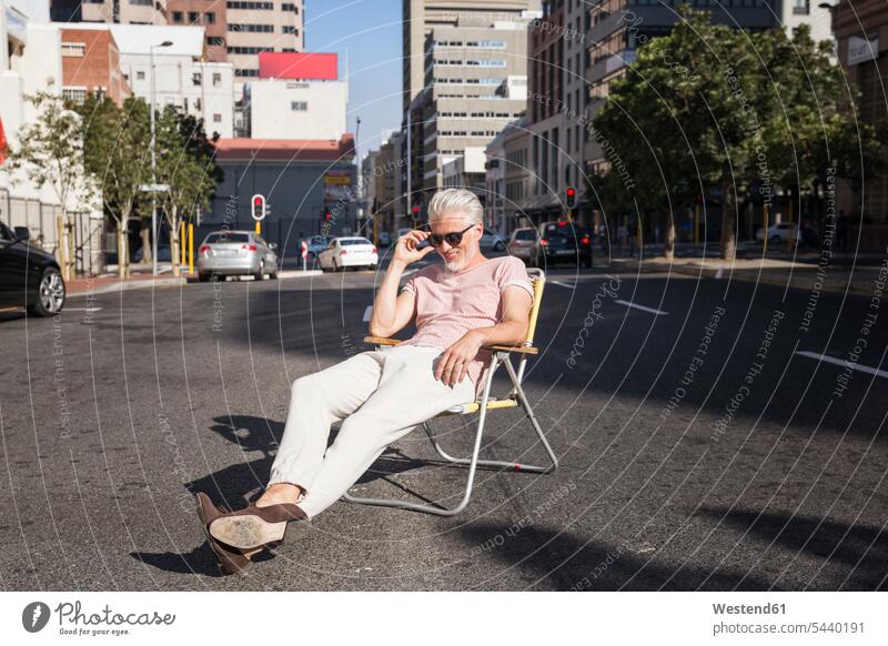 Mature man sitting on chair in the street, wearing sunglasses city town cities towns men males chairs sun glasses Pair Of Sunglasses cool attitude composed