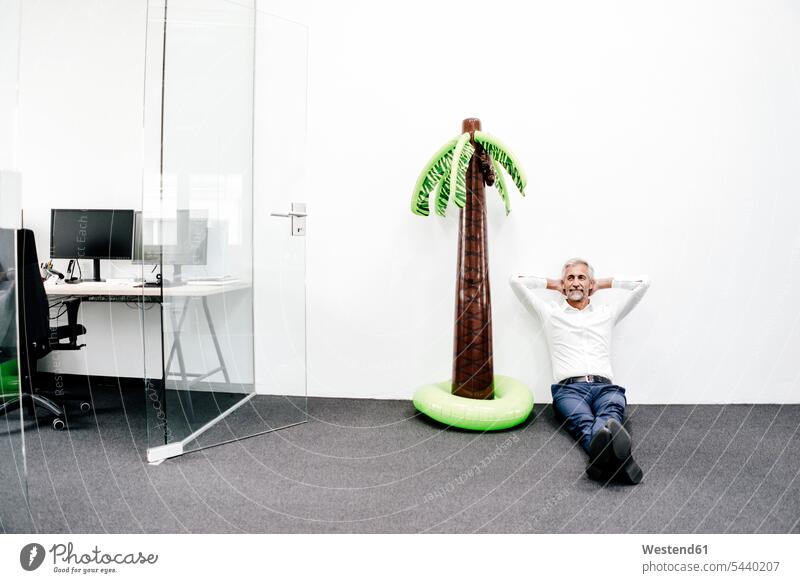 Smiling mature businessman sitting next to inflatable palm tree in office Businessman Business man Businessmen Business men smiling smile offices office room