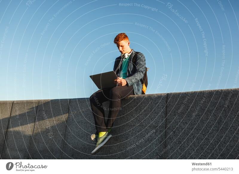 Redheaded young man sitting on wall using laptop Laptop Computers laptops notebook walls men males Seated redheaded red hair red hairs red-haired computer