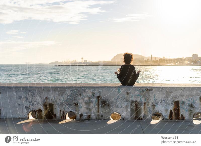 Spain, Barcelona, back view of woman sitting on wall in front of the sea meditating Seated meditate ocean females women walls water Adults grown-ups grownups