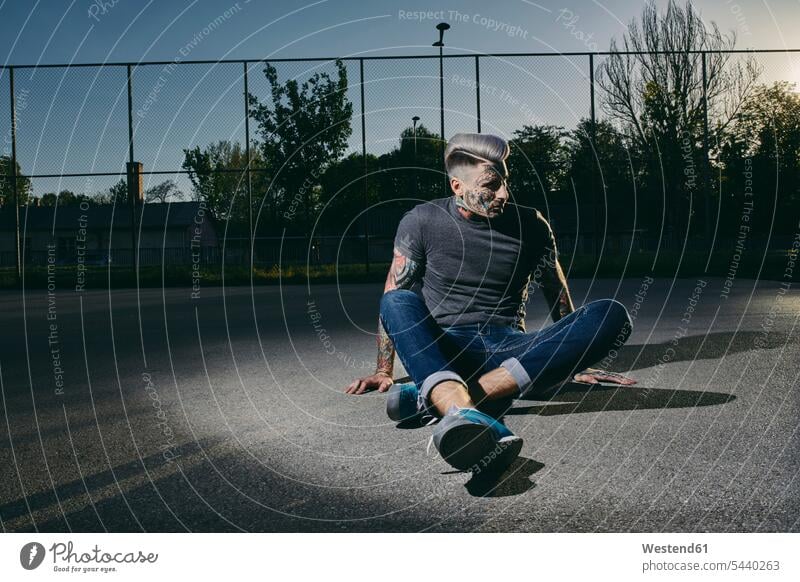 Tattooed young man sitting on basketball court at sunset sunsets sundown basketball ground men males tattoo tattoos tattooed Seated atmosphere atmospheric mood