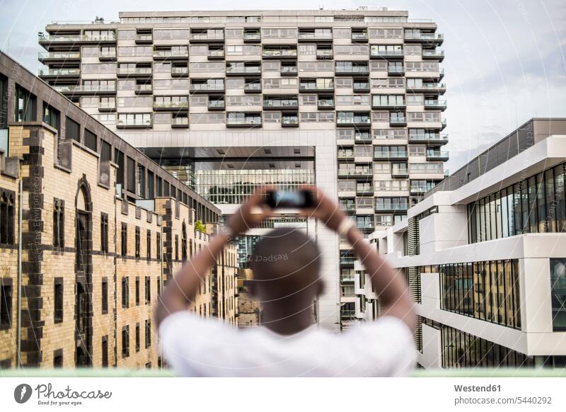 Germany, Cologne, young man taking a picture with smartphone African descent coloured black building buildings Rheinauhafen modern architecture Architecture