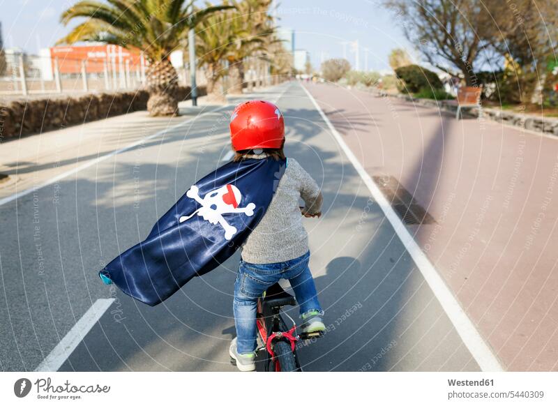 Spain, Barcelona, back view of little boy with a pirate cape riding bicycle on an empty street childhood bicycle tour bicycle tours bicycle trip bike trip paint