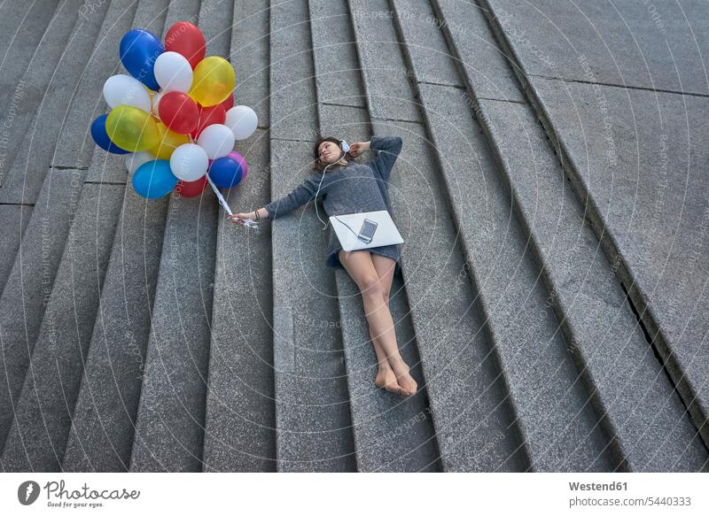 Young woman with bunch of balloons, laptop and smartphone lying on stairs listening music with headphones headset laying down lie lying down stairway Smartphone