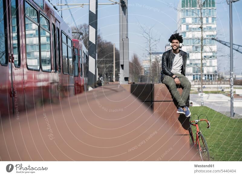 Smiling man sitting on wall listening to music next to passing train men males Seated headphones headset smiling smile Adults grown-ups grownups adult people