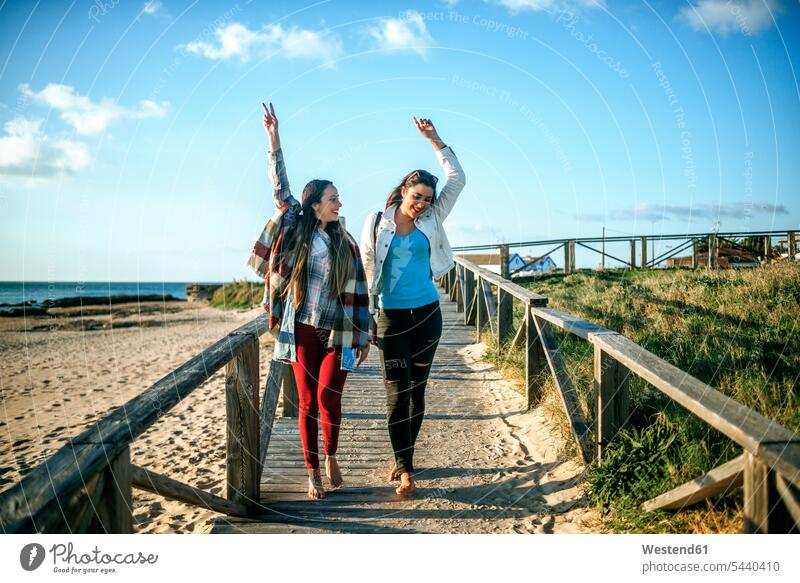 Two happy young woman walking barefoot on boardwalk female friends boardwalks wooden walkway mate friendship footbridge beach beaches going smiling smile Sea