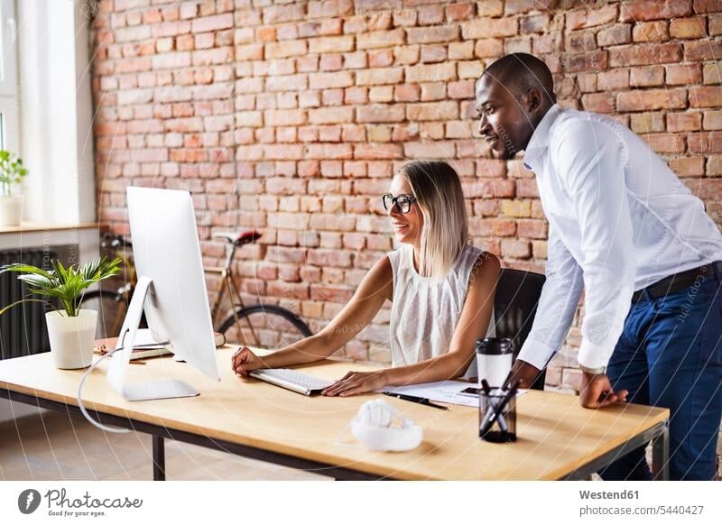 Two colleagues working together at desk in office At Work smiling smile desks offices office room office rooms talking speaking Table Tables workplace