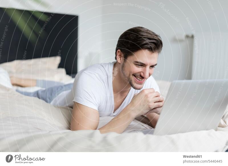 Smiling man lying on bed using laptop beds Laptop Computers laptops notebook smiling smile men males computer computers Adults grown-ups grownups adult people
