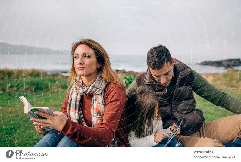 Relaxed woman with book looking aside while girl and man using tablet outdoors family families coast coastline shoreline people persons human being humans