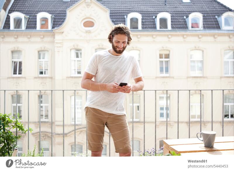 Smiling man standing on balcony looking at cell phone balconies men males Smartphone iPhone Smartphones Adults grown-ups grownups adult people persons