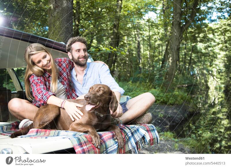 Smiling young couple with dog sitting in car at a brook in forest dogs Canine smiling smile automobile Auto cars motorcars Automobiles woods forests brooks