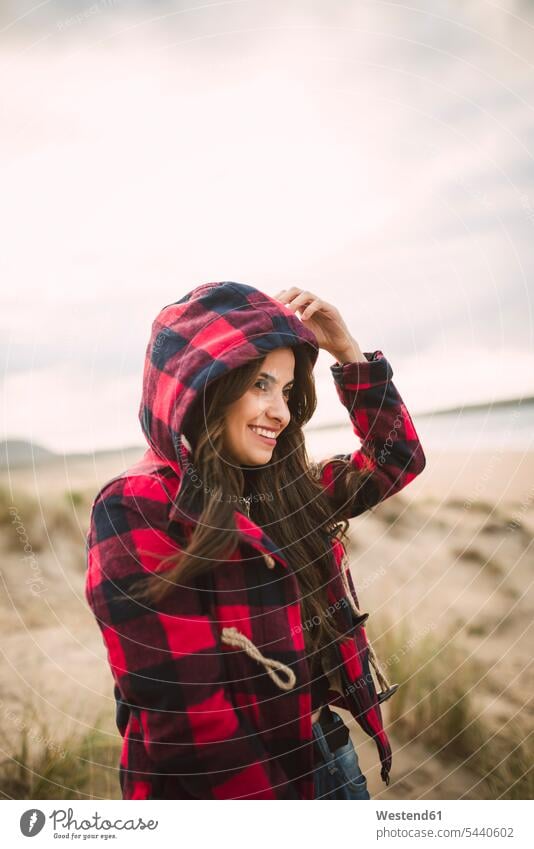 Smiling young woman with long brown hair wearing hooded jacket on the beach beaches females women Adults grown-ups grownups adult people persons human being