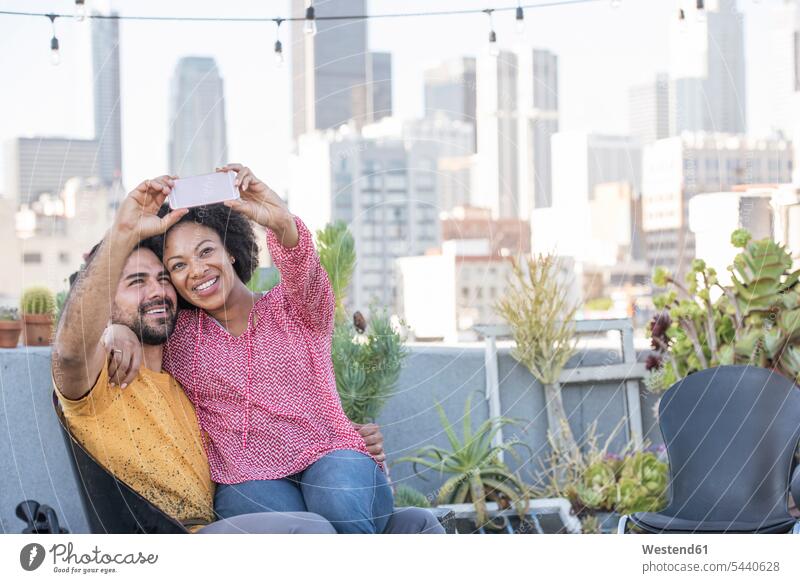 Couple sitting on rooftop terrace, taking smart phone selfies couple twosomes partnership couples photographing Seated smiling smile roof terrace deck people