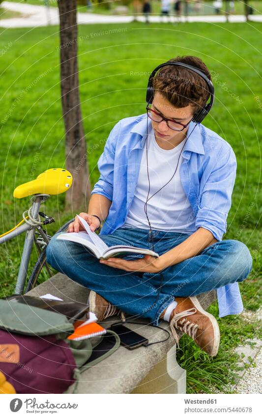 Young man with racing cycle and headphones sitting on a bench reading book headset men males books Adults grown-ups grownups adult people persons human being
