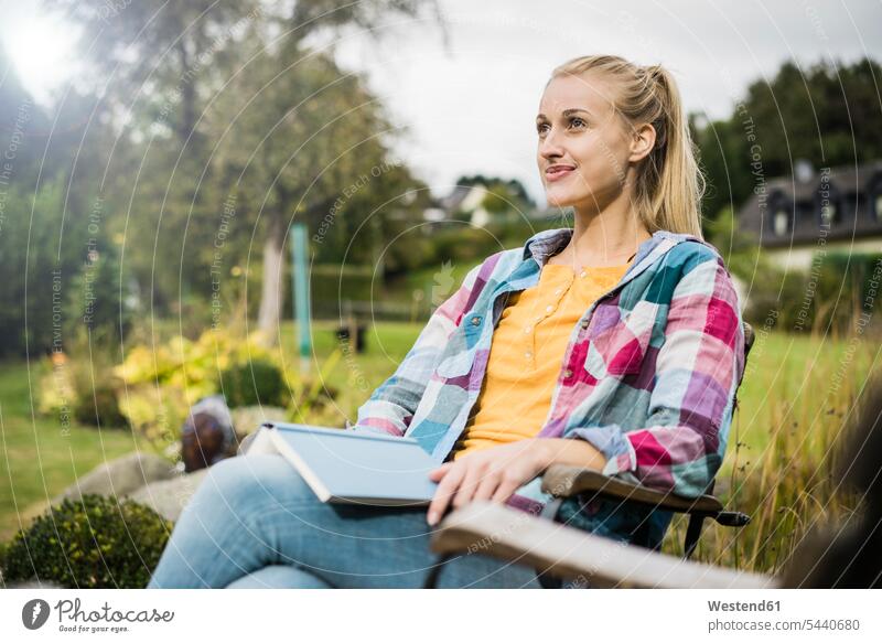 Smiling young woman with book relaxing in garden gardens books females women relaxed relaxation Adults grown-ups grownups adult people persons human being