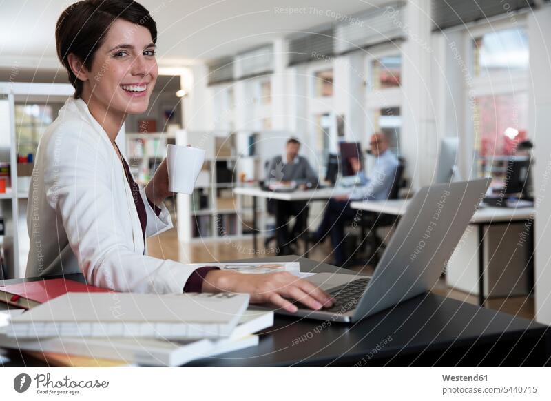 Portrait of happy woman using laptop in office with colleagues in background smiling smile businesswoman businesswomen business woman business women offices