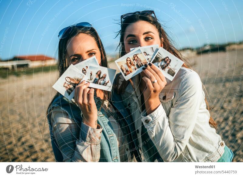 Two young women holding instant photos in front of their faces female friends beach beaches instant photography polaroid polaroids mate friendship photographs
