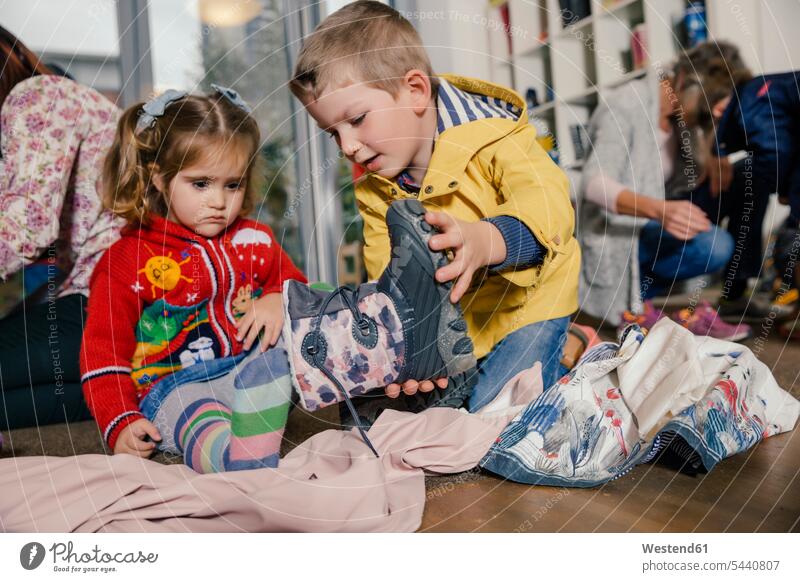 Boy helping girl putting on her boots in kindergarten child children kid kids nursery school assistance assisting Help dressing people persons human being