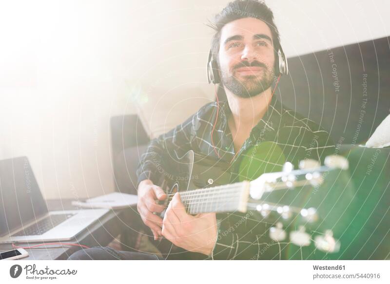 Young man at home playing guitar and wearing headphones connected to laptop guitars Laptop Computers laptops notebook smiling smile men males