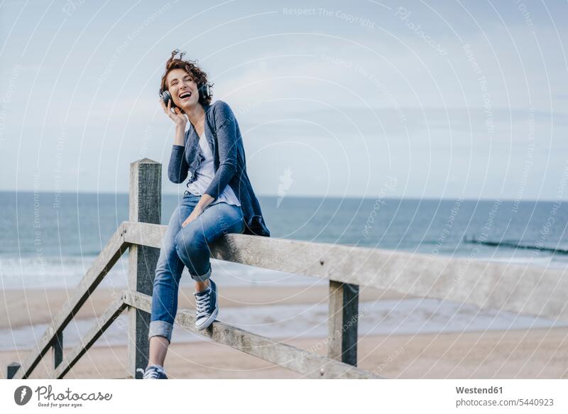 Happy woman sitting on railing at the beach listening to music beaches laughing Laughter headphones headset females women positive Emotion Feeling Feelings