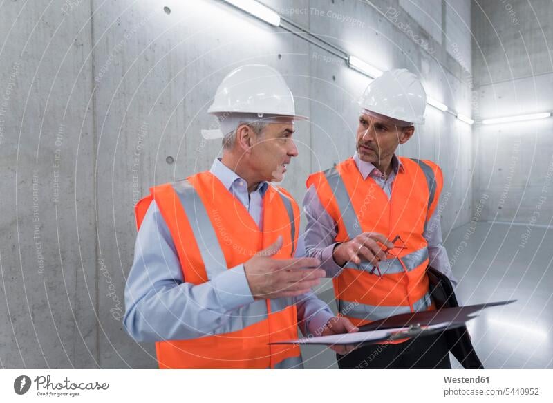 Two colleagues wearing safety vests and hard hats talking in a building working At Work man men males scrutiny scrutinizing Adults grown-ups grownups adult