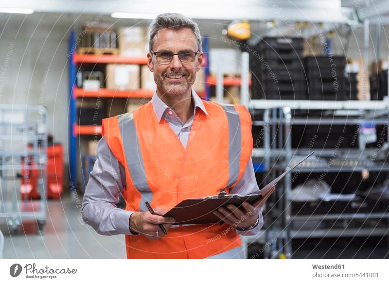 Portrait of smiling man in factory hall wearing safety vest holding clipboard working At Work smile scrutiny scrutinizing clipboards clip-board clip-boards