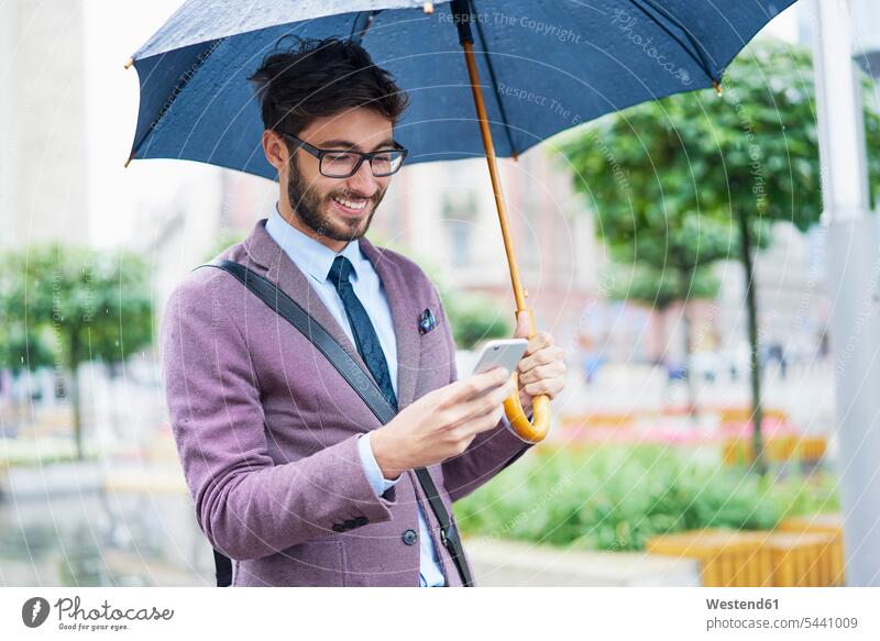 Fashionable businessman with umbrella checking his cell phone in the city fashionable Businessman Business man Businessmen Business men males umbrellas town