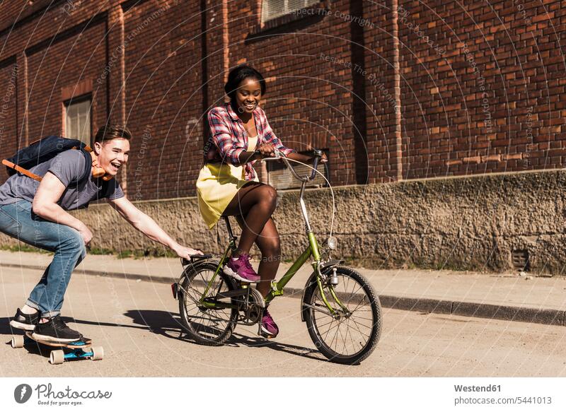 Young woman on bicycle pulling young man, standing on skateboard multicultural skateboarding Boarding cheerful gaiety Joyous glad Cheerfulness exhilaration