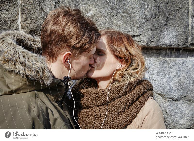 Young couple sharing earbuds kissing at stone wall kisses twosomes partnership couples earphones ear phone ear phones people persons human being humans
