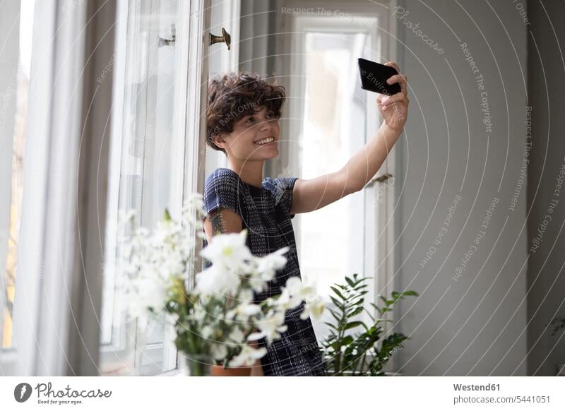 Smiling young woman standing in front of window taking selfie with smartphone Selfie Selfies photographing females women Adults grown-ups grownups adult people